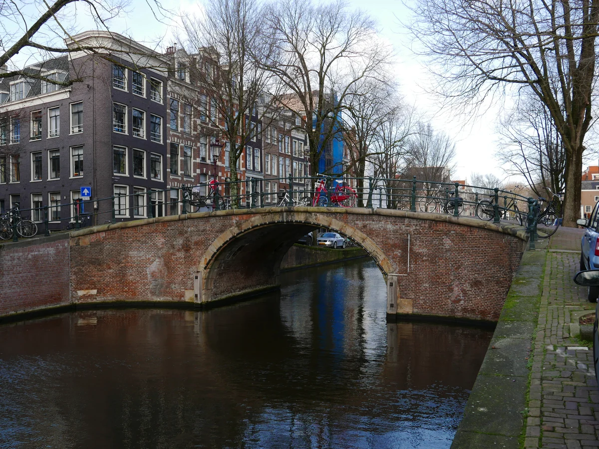 A brick arch bridge over an Amsterdam canal with gabled houses on the left and bicycles parked on the bridge, reflected in the still water.