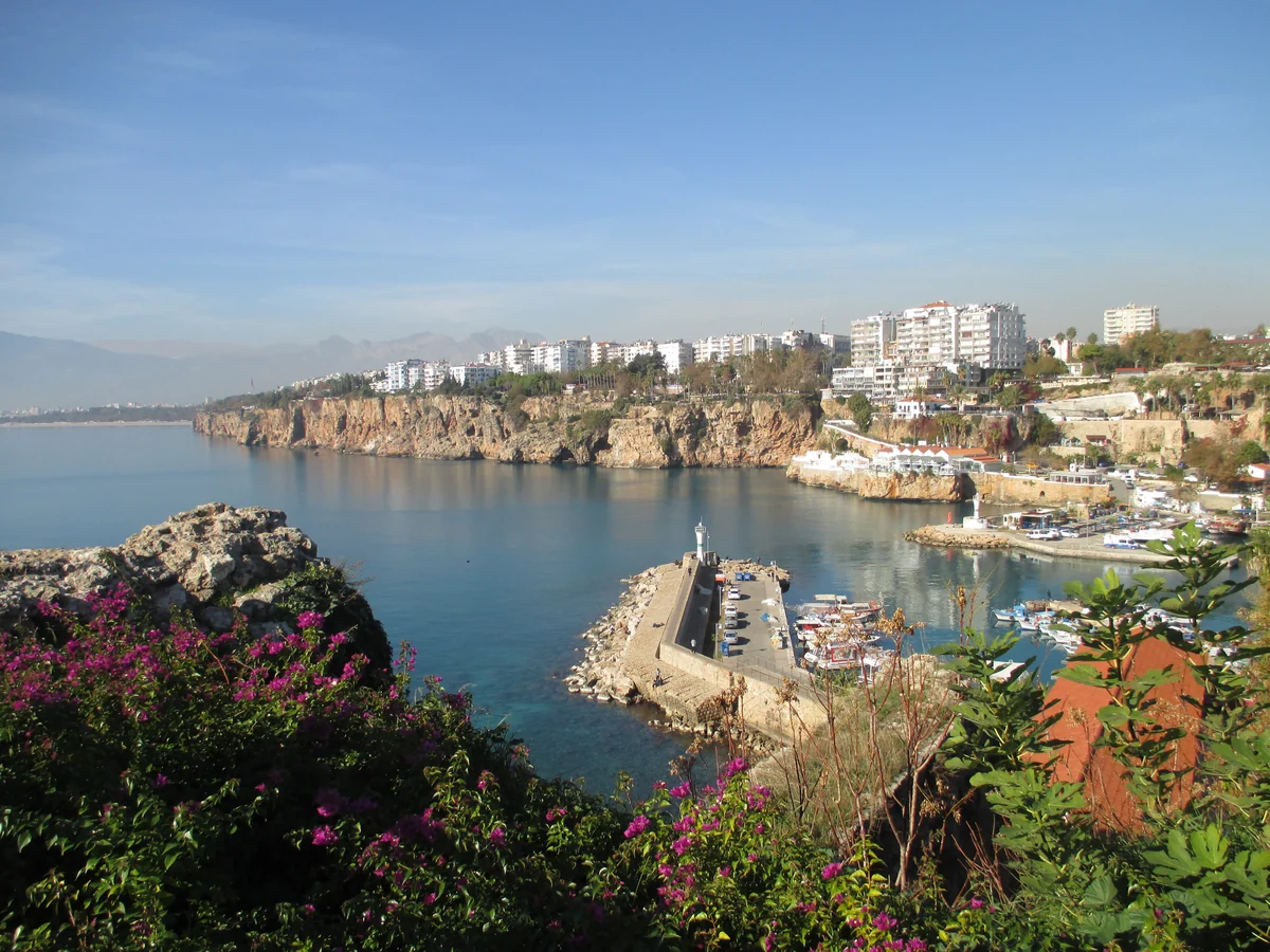 A high-angle view of the Antalya old city harbour at the foot of a steep cliff topped with Kaleiçi buildings, with the Taurus Mountains in the background under a late afternoon sky.