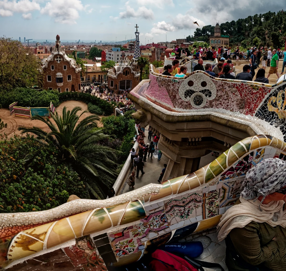 A vibrant documentary photograph of the serpentine mosaic bench at Park Güell, with the iconic gingerbread-style gatehouses and the city of Barcelona in the background.