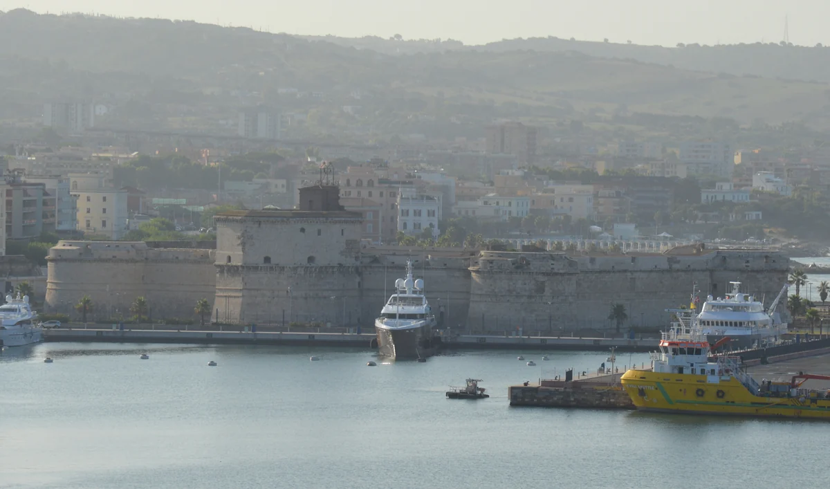 A morning view of the historic Fort Michelangelo in Civitavecchia, with its stone bastions shrouded in a thick harbor haze and several ships moored in the surrounding water.