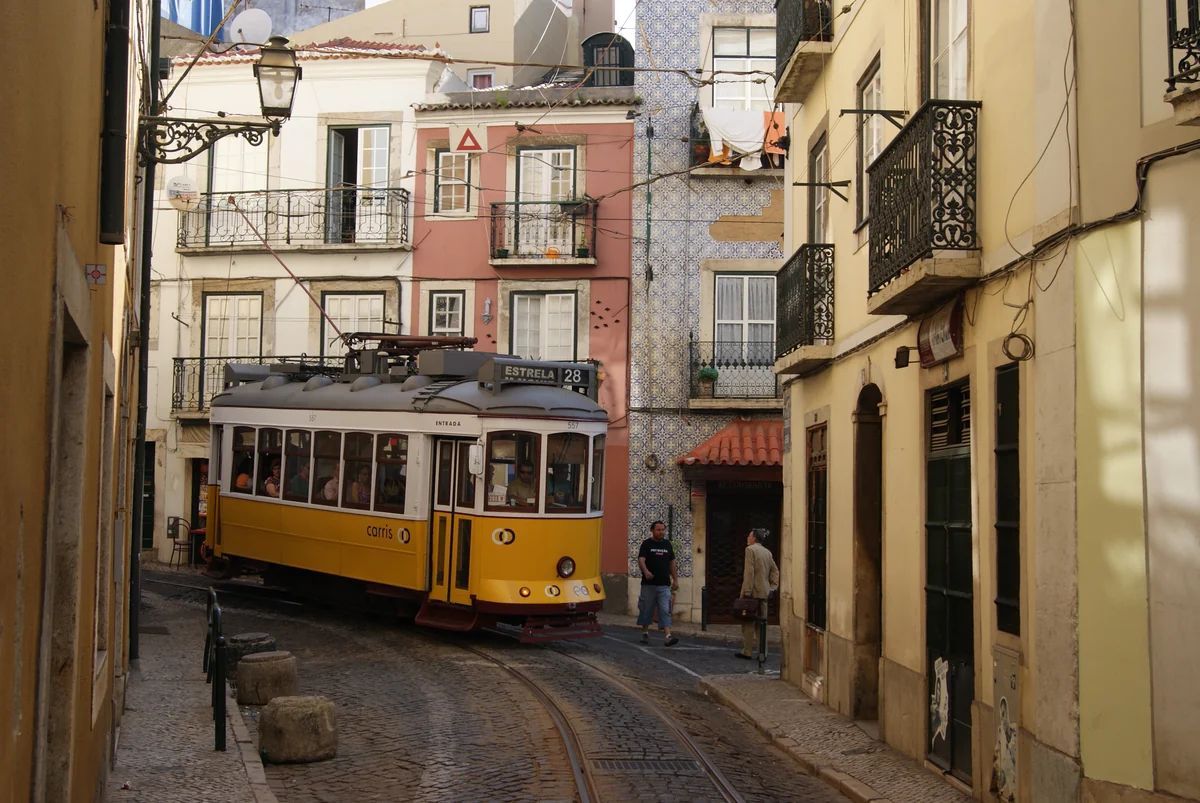 A vintage yellow tram 28 climbs a narrow cobblestone street in Lisbon's Alfama district, passing a building with a traditional azulejo tile façade with the Tejo river visible in the distant background.