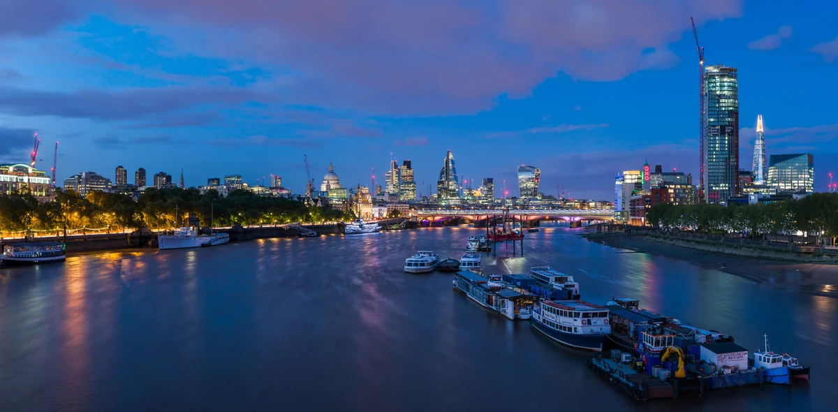 A wide panorama of the London skyline at dusk featuring the curve of the River Thames, Blackfriars Bridge, and the illuminated dome of St Paul's Cathedral.