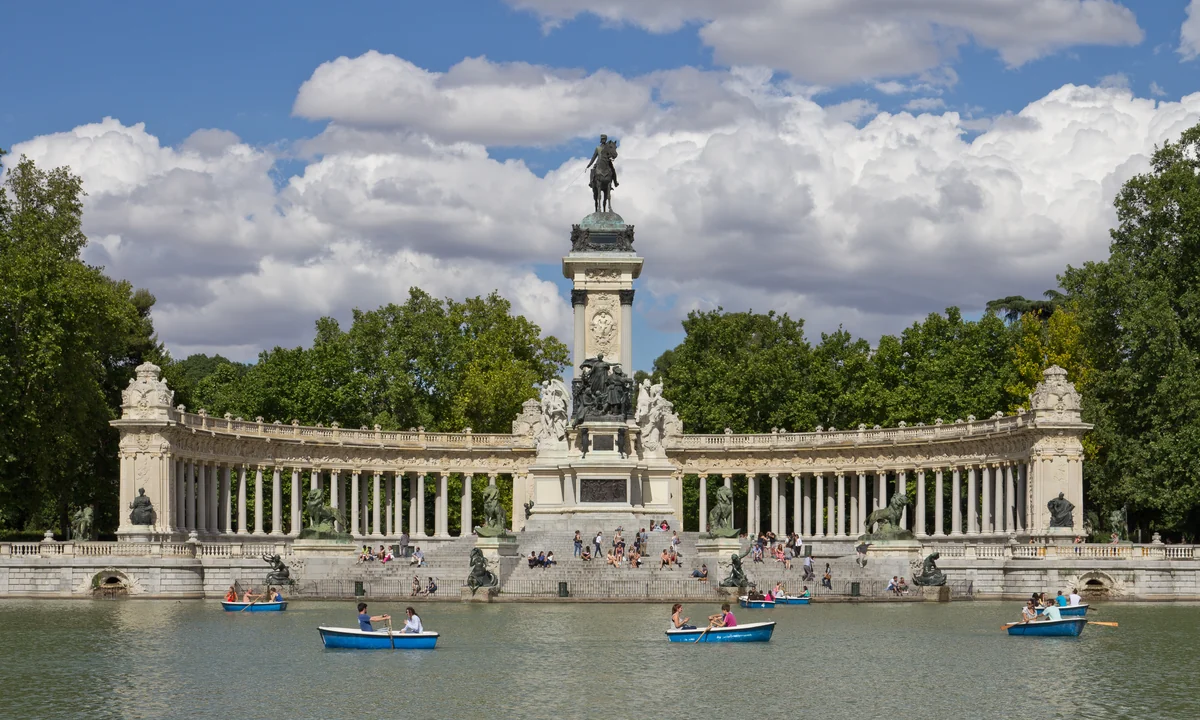 A panoramic view of the semicircular colonnade and equestrian statue of the Monument to Alfonso XII in Madrid's Retiro Park, reflected in the calm waters of the park's lake with city rooftops visible in the distance.