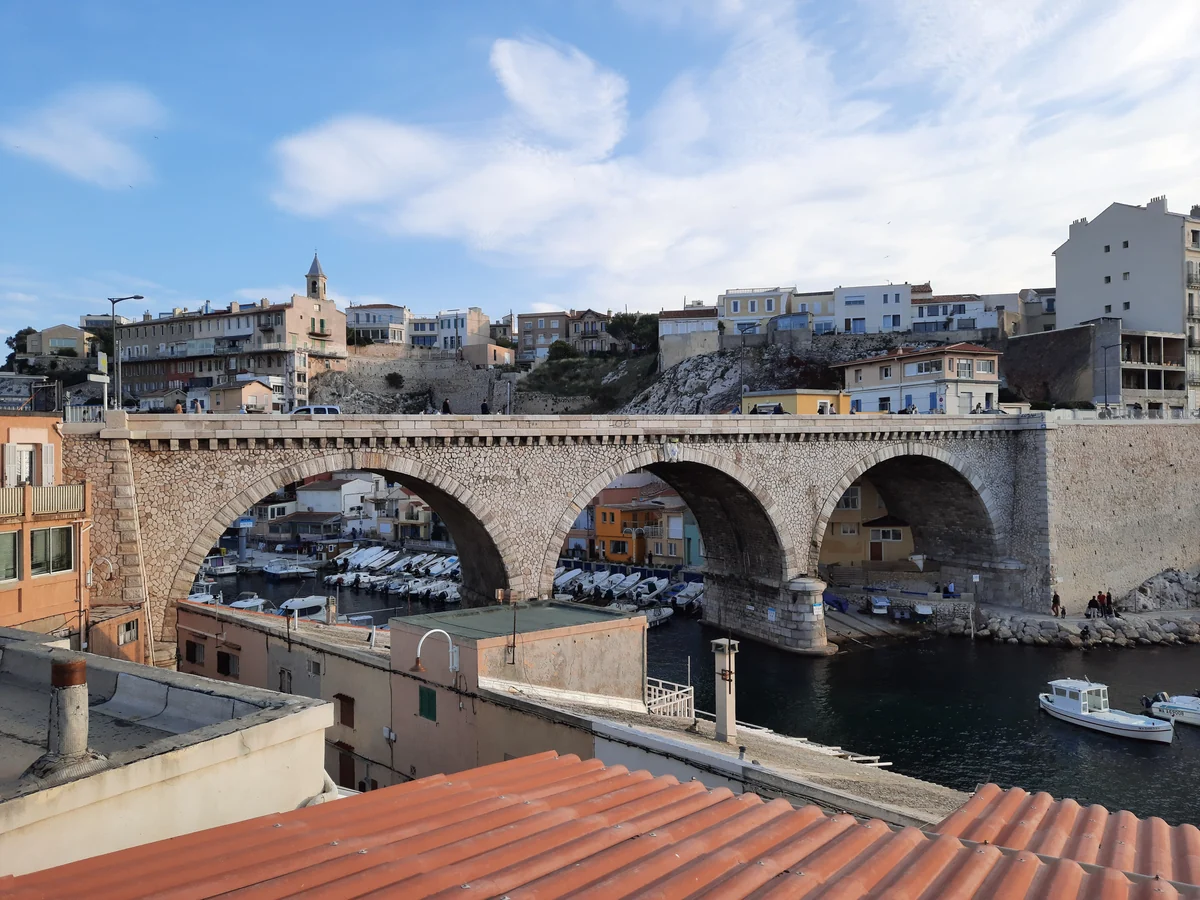 A view of the Vallon des Auffes fishing port in Marseille with traditional boats moored under a stone bridge and the Notre-Dame de la Garde basilica on a hill in the distance.
