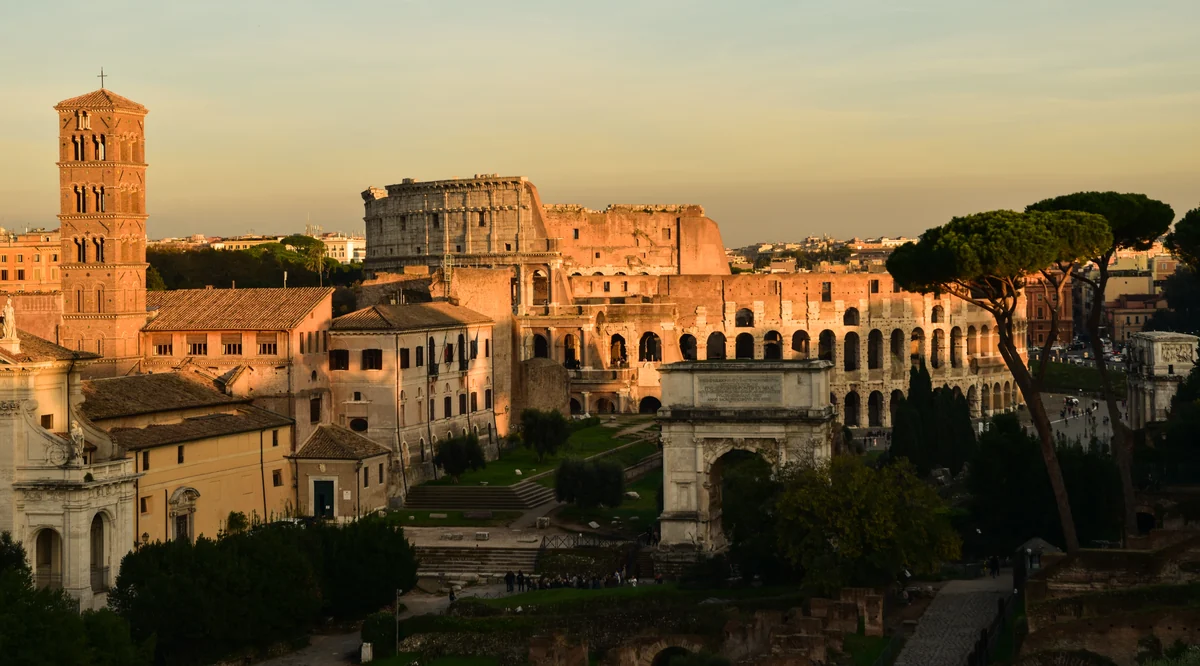 The ancient stone arches of the Colosseum in Rome are illuminated by the warm glow of a sunset, with a prominent umbrella pine tree standing to the right and domes visible in the distant city skyline.