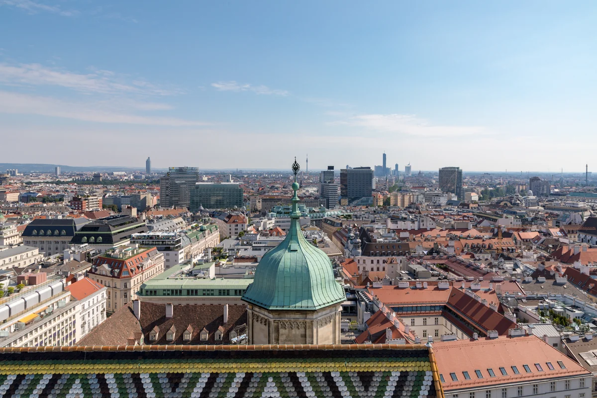A high-angle view of the steep, chevron-patterned colorful roof tiles of St. Stephen's Cathedral in Vienna, with a green-domed tower in the foreground and the historic city skyline in the background.